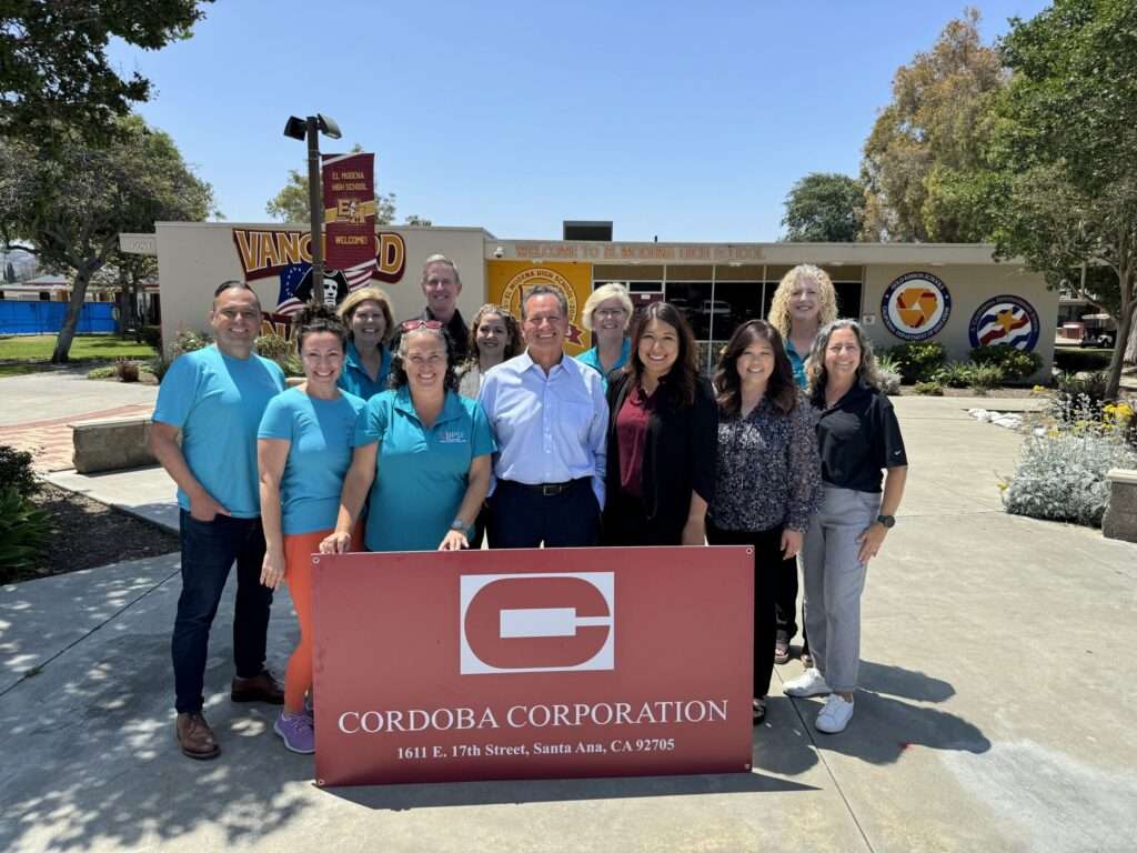 A group of people stands outside El Modena High School in front of a Cordoba Corporation sign. The group includes individuals wearing turquoise shirts with the Cordoba Corporation logo and others in business casual attire, smiling for the photo. The school building and signage are visible in the background on a sunny day.
