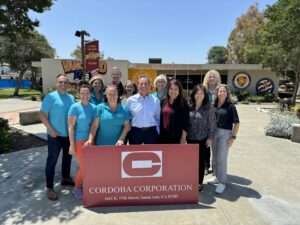 A group of people stands outside El Modena High School in front of a Cordoba Corporation sign. The group includes individuals wearing turquoise shirts with the Cordoba Corporation logo and others in business casual attire, smiling for the photo. The school building and signage are visible in the background on a sunny day.