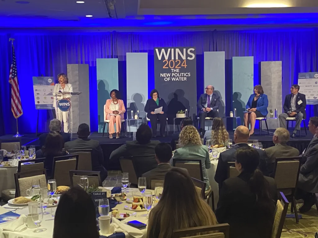 A speaker addresses the audience at a conference titled "WINS 2024: The New Politics of Water." The stage features a panel of five seated individuals, and the event is set in a large banquet hall with attendees seated at round tables. The room is illuminated with blue lighting, and the backdrop includes event branding and sponsor logos.