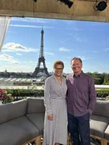 A couple smiling and posing in front of the Eiffel Tower in Paris, France. The woman is wearing a black and white striped dress, and the man is in a purple shirt. They are standing on an outdoor patio with a clear view of the iconic tower against a bright blue sky.