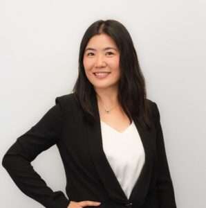 This image shows a woman smiling confidently at the camera. She has medium-length dark hair, is wearing a black blazer over a white top, and has a necklace with a small pendant. She stands against a plain light-colored background with her hand on her hip.