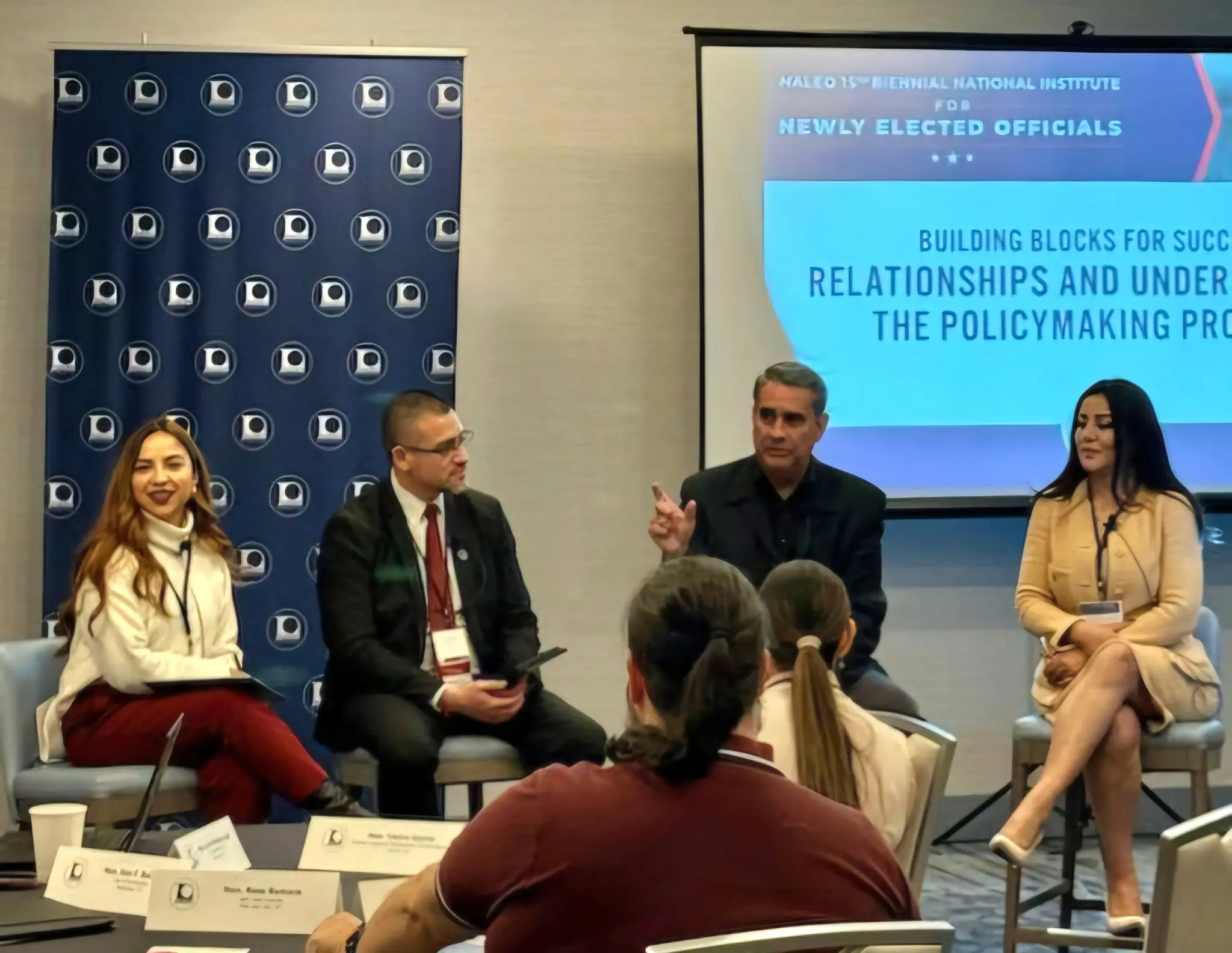 This image shows a panel discussion at an event, with four speakers seated in front of an audience. Behind them, a banner with a logo and a presentation screen displaying the title "Building Blocks for Success: Relationships and Understanding the Policymaking Process" is visible. The scene conveys a professional and educational atmosphere.